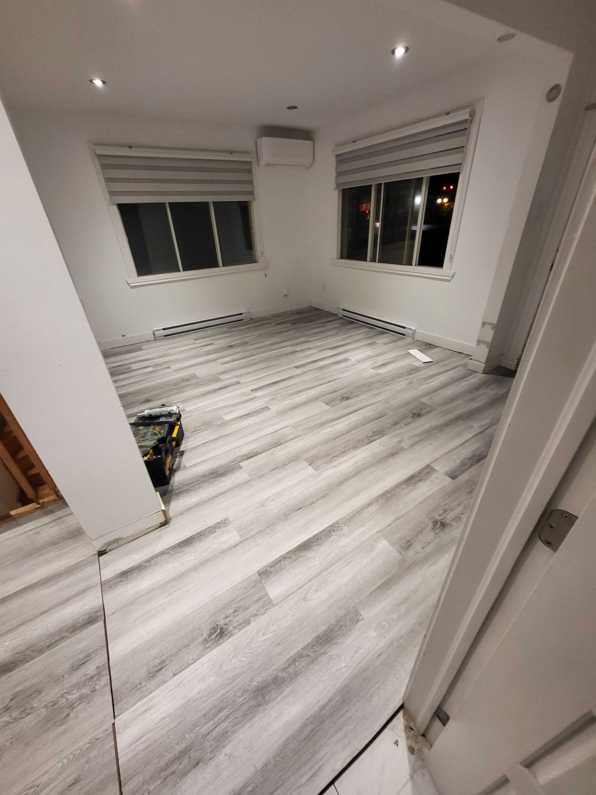 Pièce moderne vide avec parquet gris clair, murs blancs et deux fenêtres aux stores ouverts.