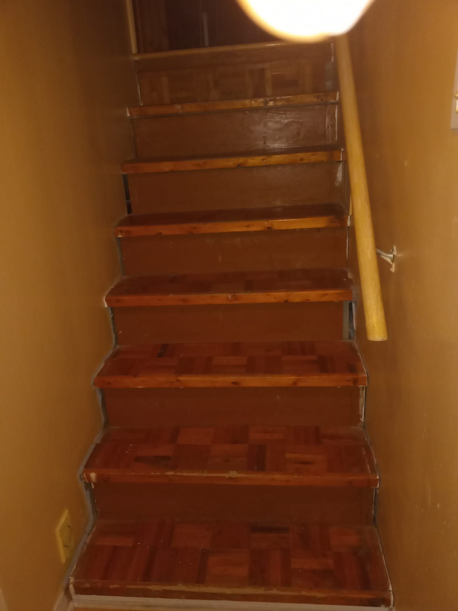 Un étroit escalier en bois dans un couloir sombre, éclairé d'une lumière chaude, avec une rampe jaune sur la droite.