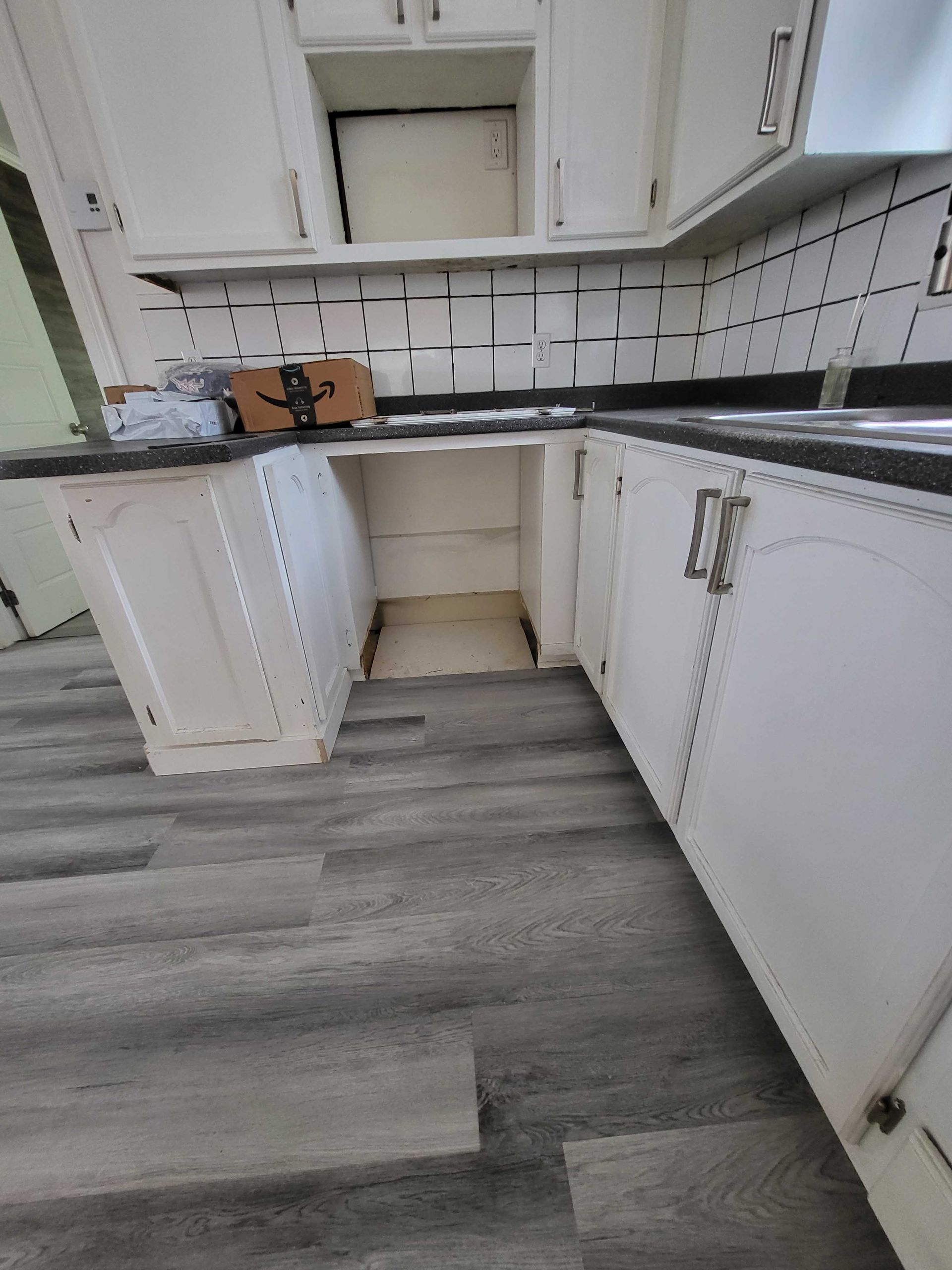Cuisine blanche avec placards, comptoirs noirs, crédence en carrelage blanc et sol gris imitation bois.