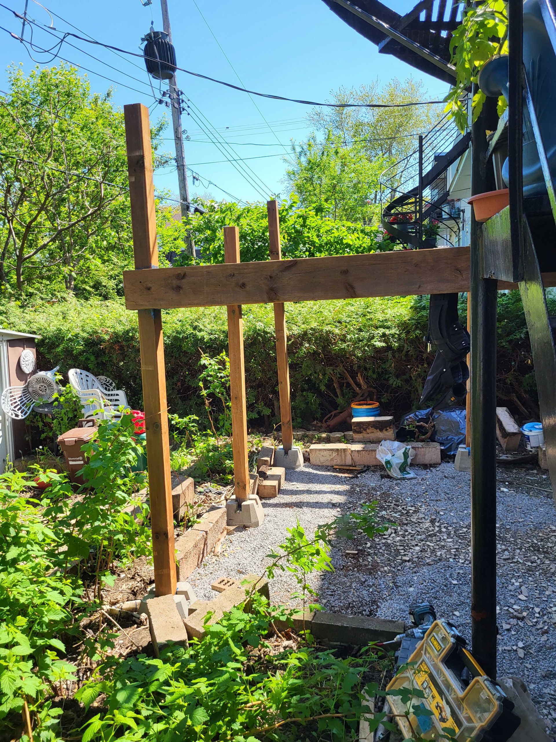 Poteaux en bois dressés dans un jardin de gravier en construction, avec des plantes vertes et un ciel bleu en arrière-plan.