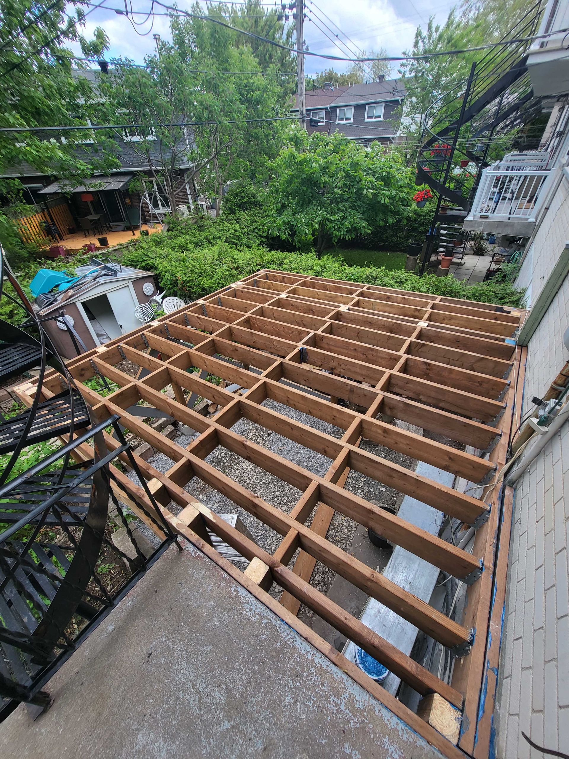 Cour arrière avec charpente de terrasse en bois apparente en construction, entourée d'arbres et de maisons en briques