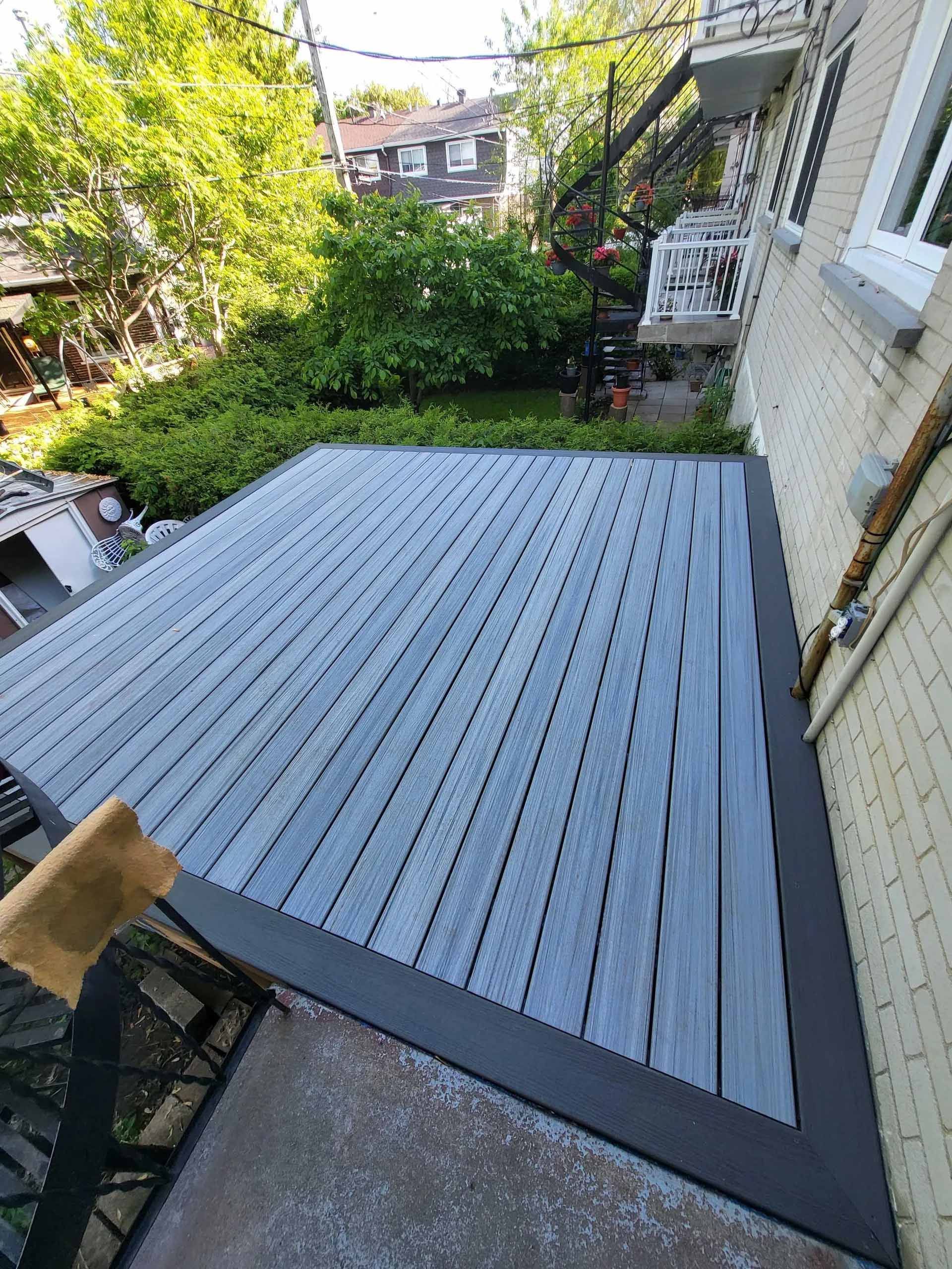 Terrasse grise surélevée dans une cour arrière attenante à une maison, entourée d'arbres et de verdure.