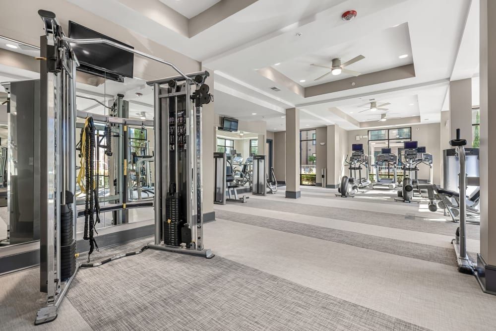 Modern gym interior with various exercise machines, including a cable machine in the foreground, treadmills, elliptical trainers, and stationary bikes. The room has bright lighting, large mirrors on the left wall, and a view of an outdoor patio area through glass doors in the background at Marq at Crabtree in Raleigh, NC.