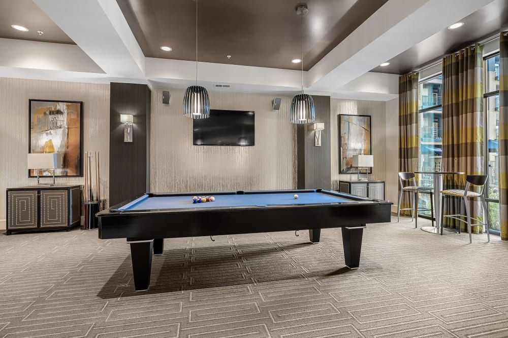 A spacious and modern recreational room featuring a blue-felt pool table with racked balls, cues lined against a wall, and an adjacent cabinet. Elegant pendant lights hang above, complementing the contemporary decor. To the right, a high-top table with stools is positioned beside floor-to-ceiling windows draped with striped curtains, offering a view of an urban landscape outside. The room has patterned carpet flooring, a mounted television, and artistic wall paintings, creating a cozy yet upscale ambiance at Marq at Crabtree in Raleigh, NC.
