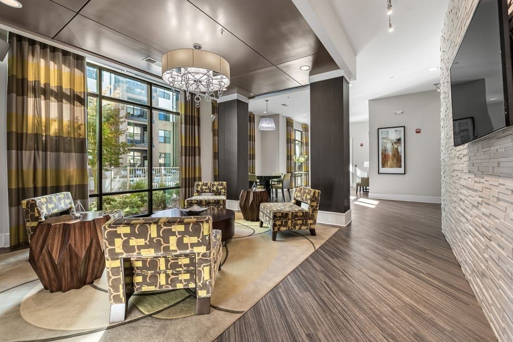 Modern and stylish lounge area with patterned armchairs and wooden tables on a beige rug, a large floor-to-ceiling window with striped curtains looking out to an urban view, elegant light fixtures, a framed artwork on the wall, and a textural white stone column to the right at Marq at Crabtree in Raleigh, NC.