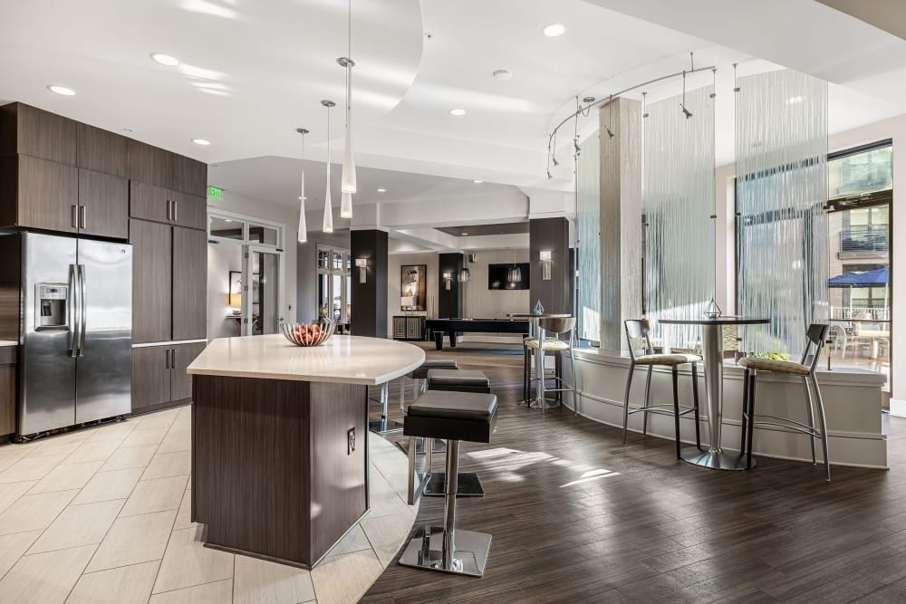 Modern and stylish kitchen and dining area in clubhouse with dark wood flooring, featuring a central island with a curved countertop, high stools, stainless steel appliances, pendant lights, and decorative vertical glass dividers that partially separate the space from the seating area with bar stools in the background at Marq at Crabtree in Raleigh, NC.