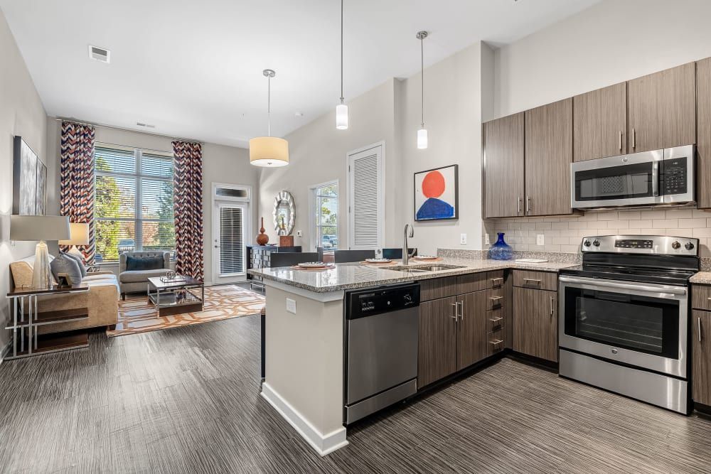 Modern apartment interior featuring an open-plan kitchen with granite countertops and stainless steel appliances, and a living area with stylish furniture, hardwood floors, and ample natural light from large windows. Decor includes vibrant curtains and a contemporary art piece at Marq at Crabtree in Raleigh, NC.