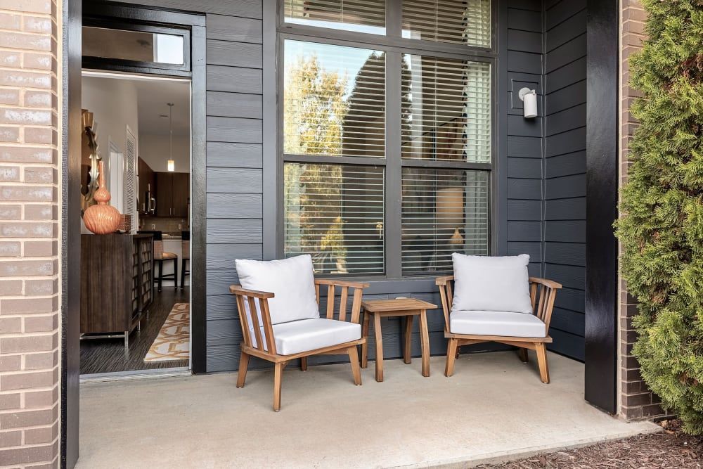 A cozy and welcoming porch featuring two wooden armchairs with white cushions, a matching wooden side table, and a view into a warmly lit interior with modern furnishings. The dark exterior walls contrast with the brick accents while a lush green shrub adds a touch of nature at Marq at Crabtree in Raleigh, NC.