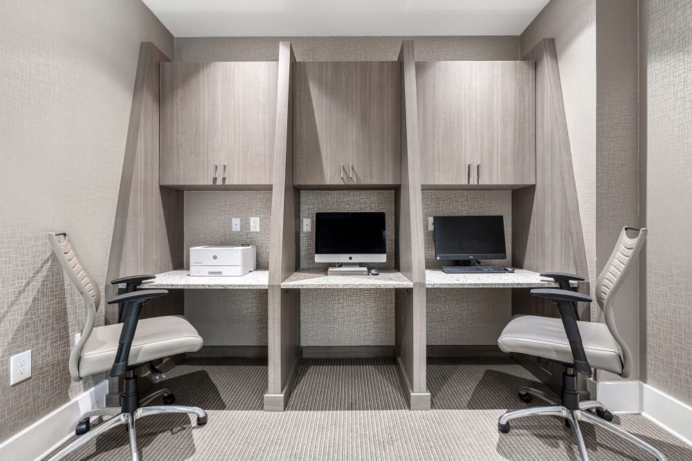 Modern dual workstation in a business center with two desktop computers on a shared counter, ergonomic chairs, and built-in light wood shelving units against a textured wallpapered wall at Marq at Crabtree in Raleigh, NC.