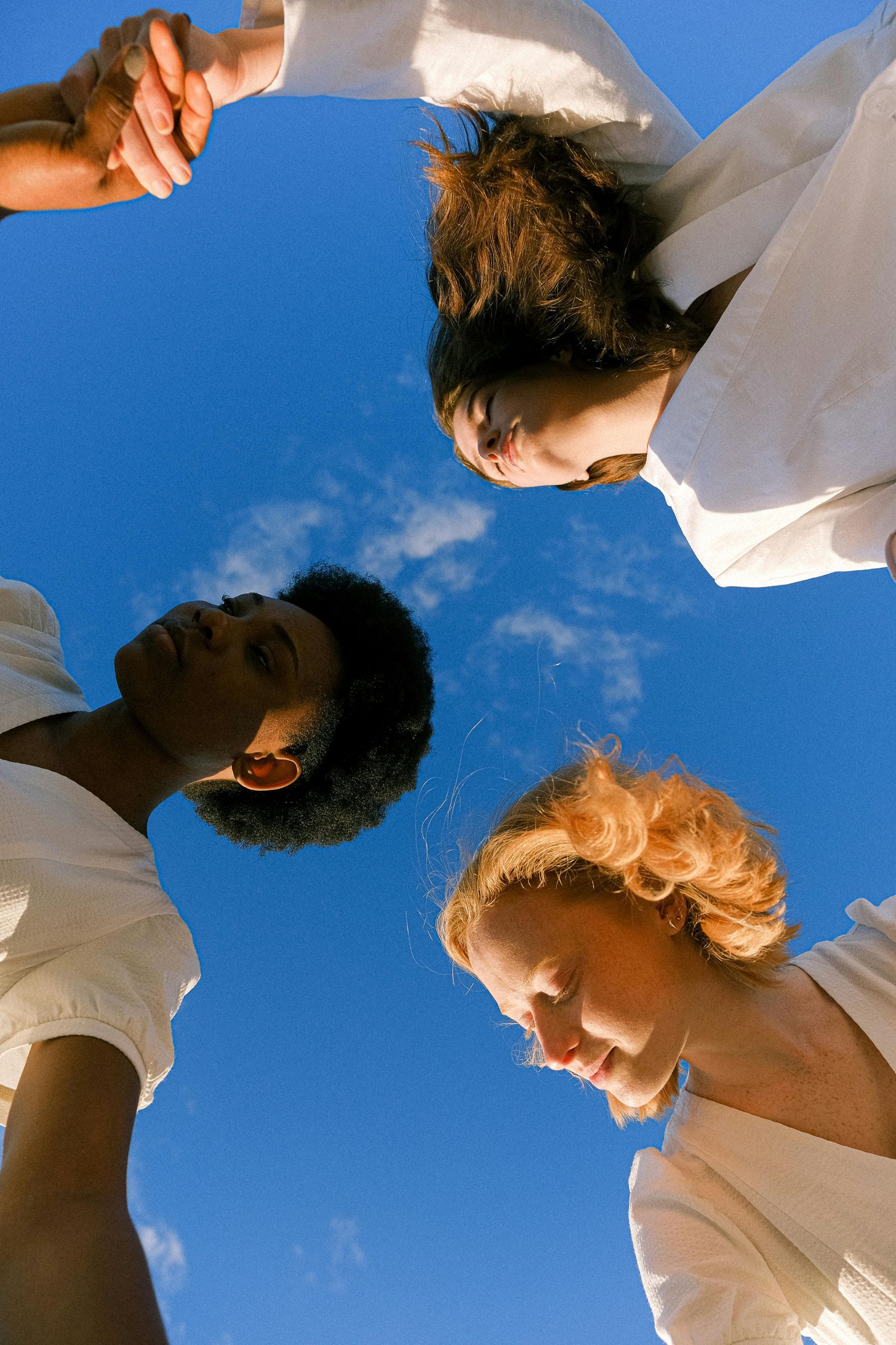 Three people wearing white tops look downward, their arms joined in a circle against a bright blue sky.