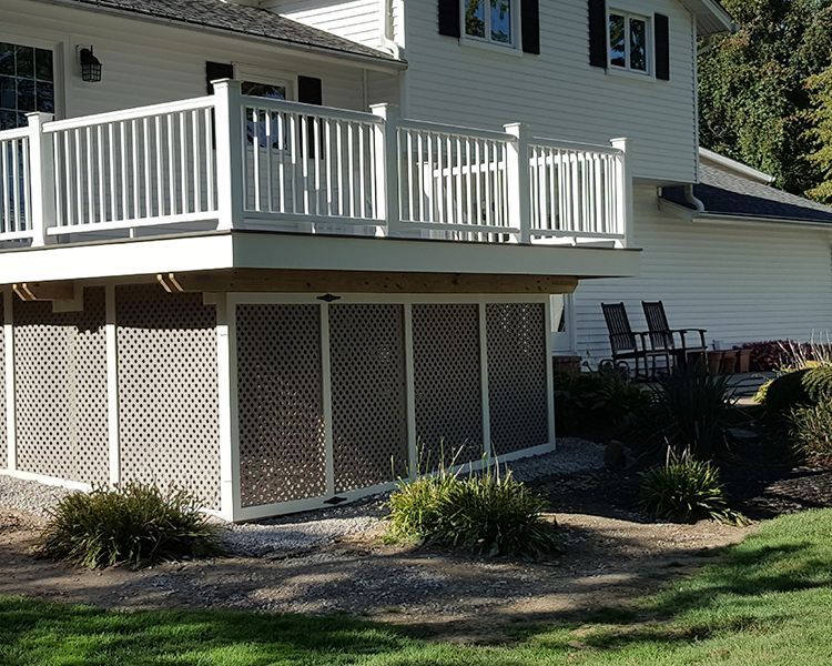 A white house with a raised wooden deck featuring white railings and lattice screening underneath in a sunny yard.
