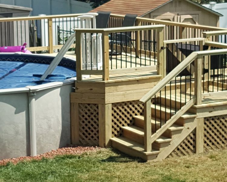 A wooden deck with stairs and lattice skirting built next to an above-ground swimming pool with a blue cover.