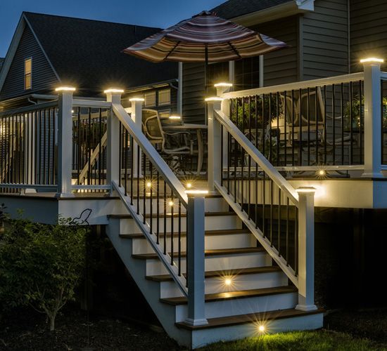 A wooden deck with white railings and illuminated post caps, featuring stairs at night with integrated step lighting.