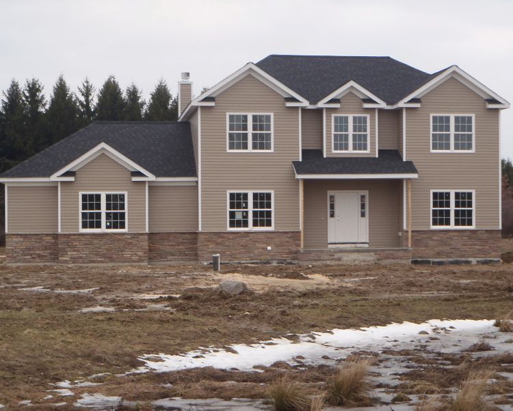 A two-story house with tan siding, a stone veneer base, and a dark roof, set on a grassy lot with patches of snow.