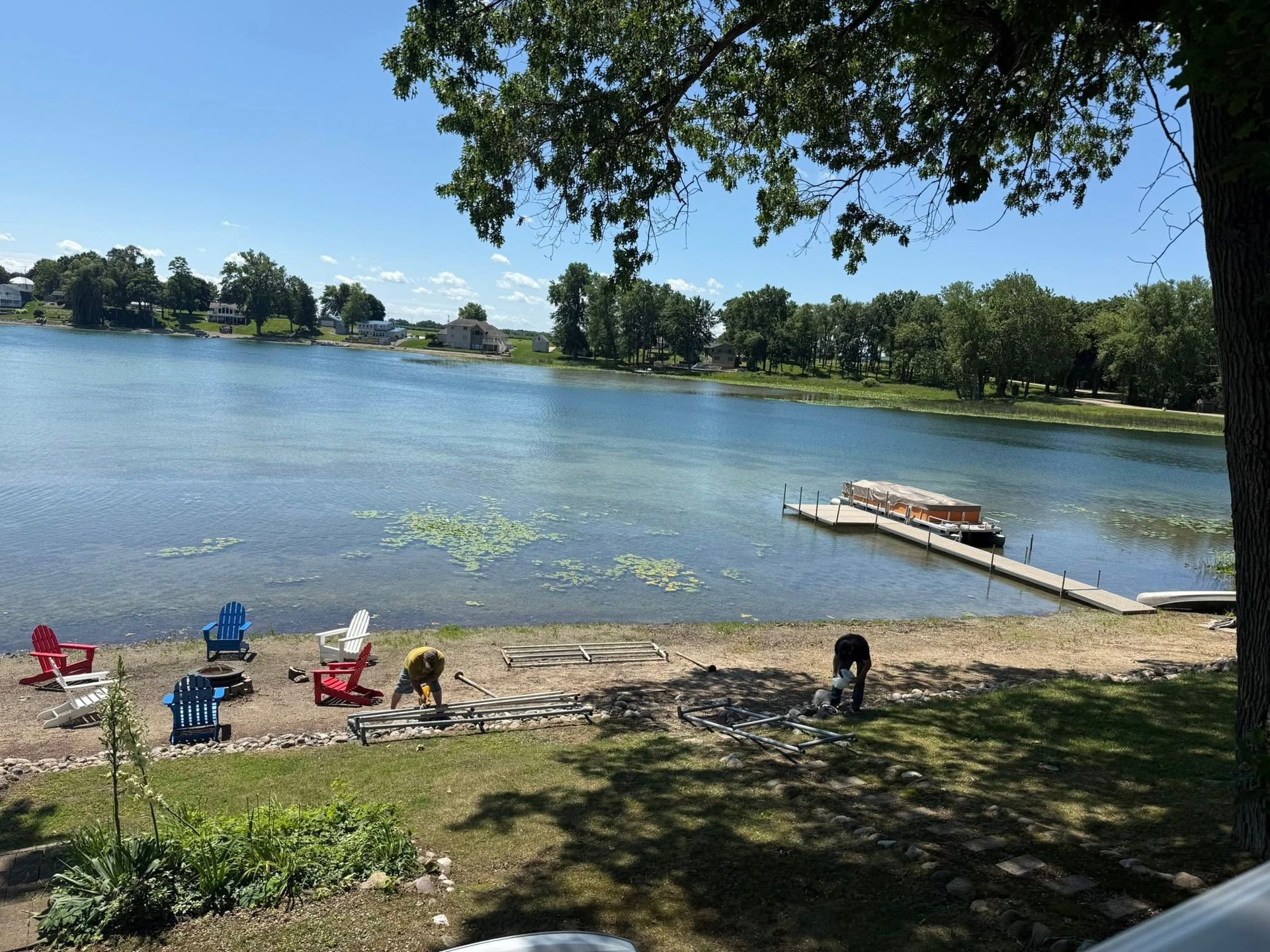 Sunny lakeside beach with chairs, people relaxing, and a small dock on calm water