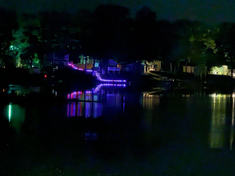 Nighttime riverside scene with neon purple and green lights reflecting on calm water, trees silhouetted above