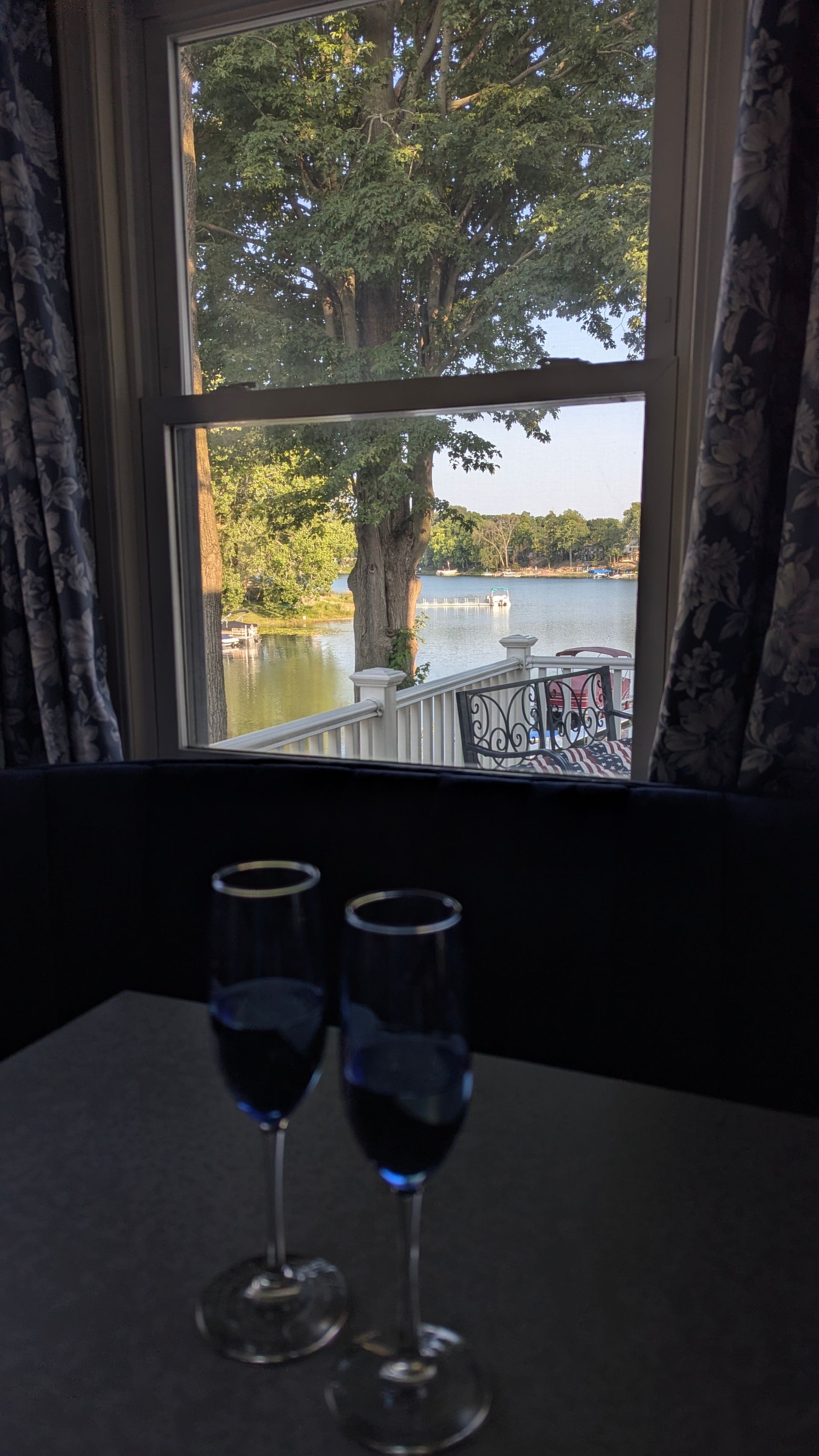 Two wine glasses on a table by a window overlooking a lakeside view.