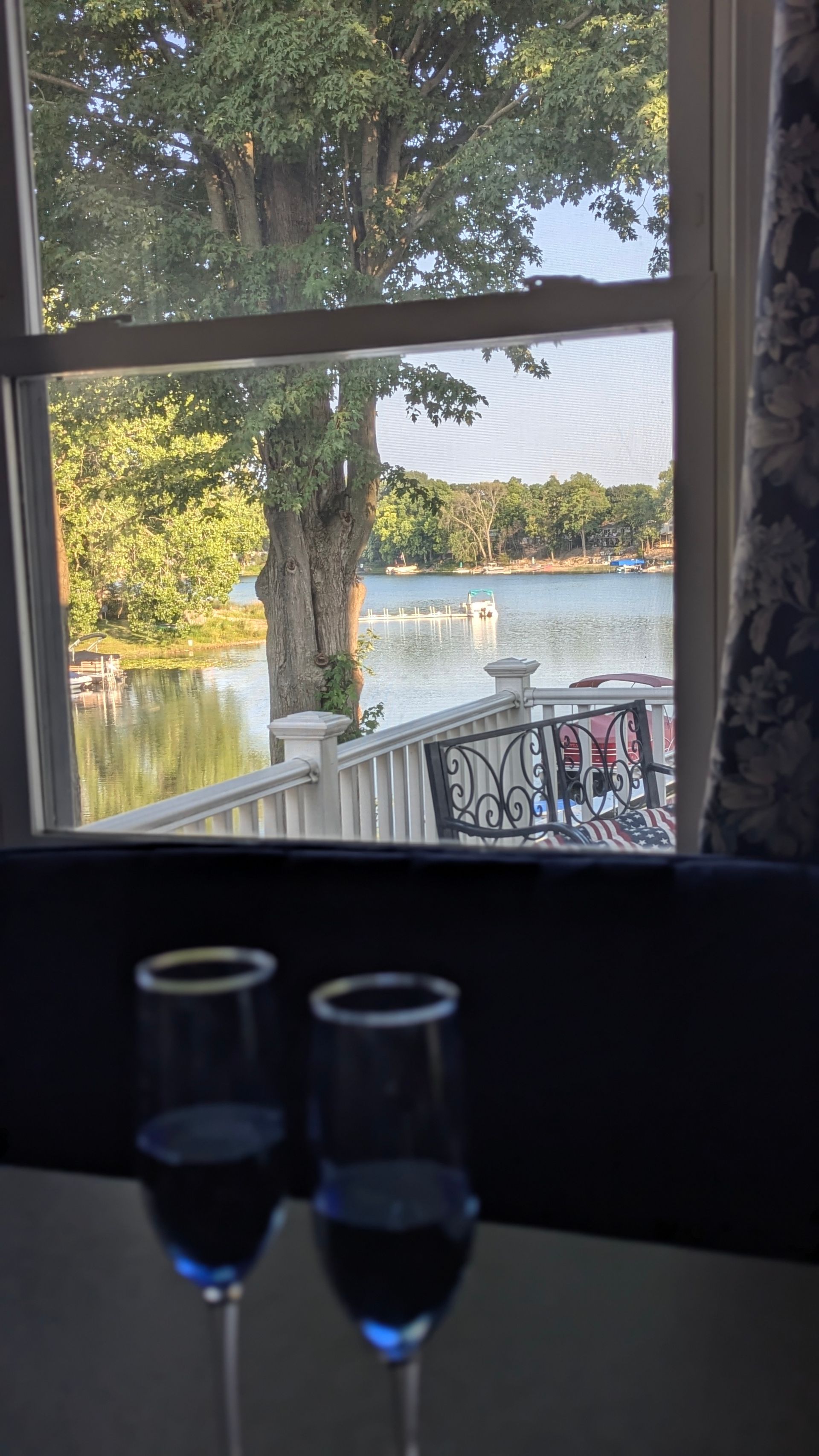 Two wine glasses on a table by a window overlooking a lake and trees