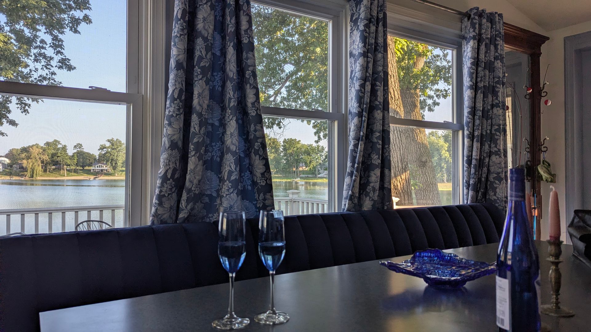 Dining table by large windows overlooking a lake, with blue glasses and dark drapes.
