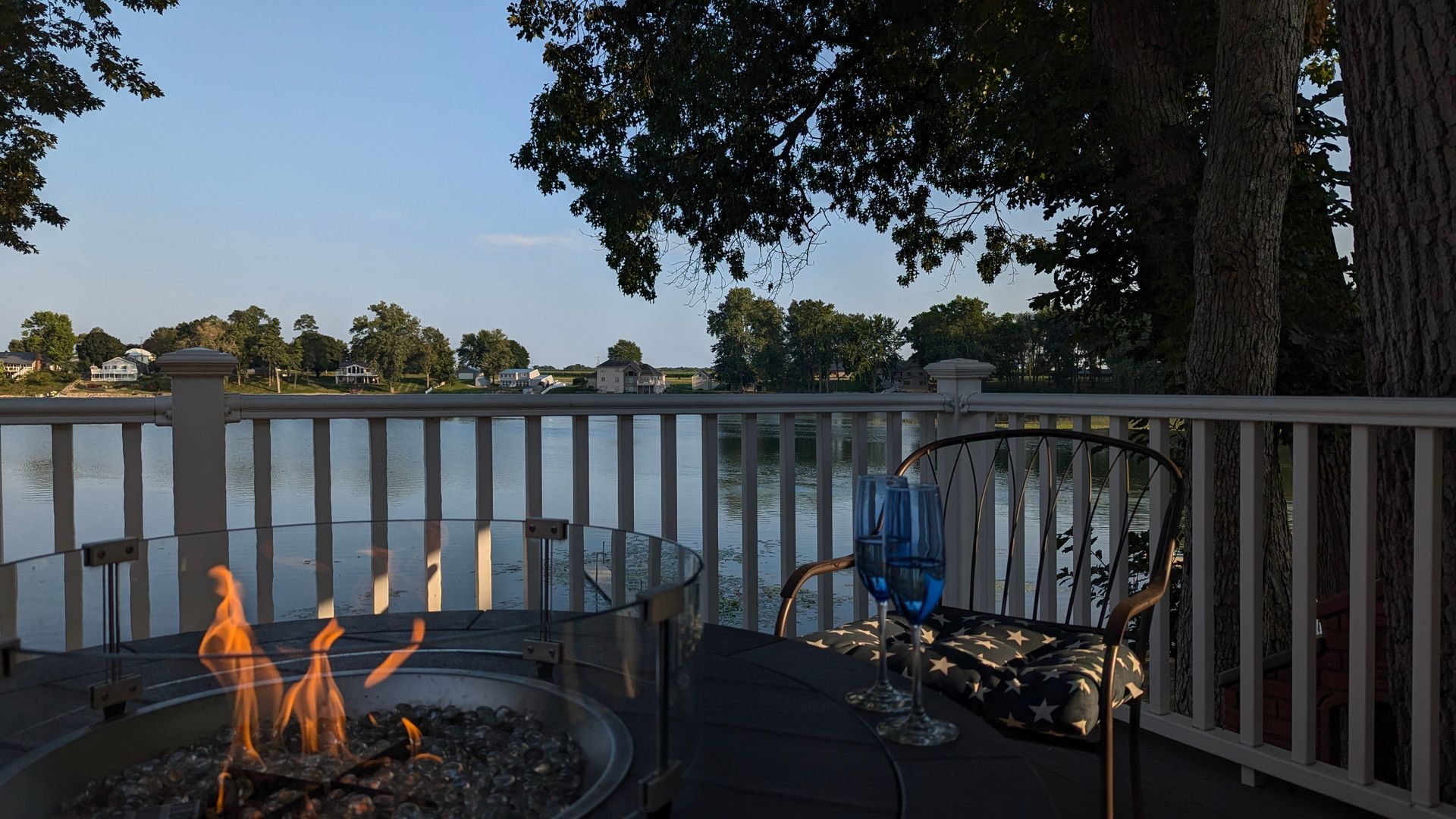 Lakeside deck at dusk with a fire pit, two chairs, white railing, and trees reflecting on the water.