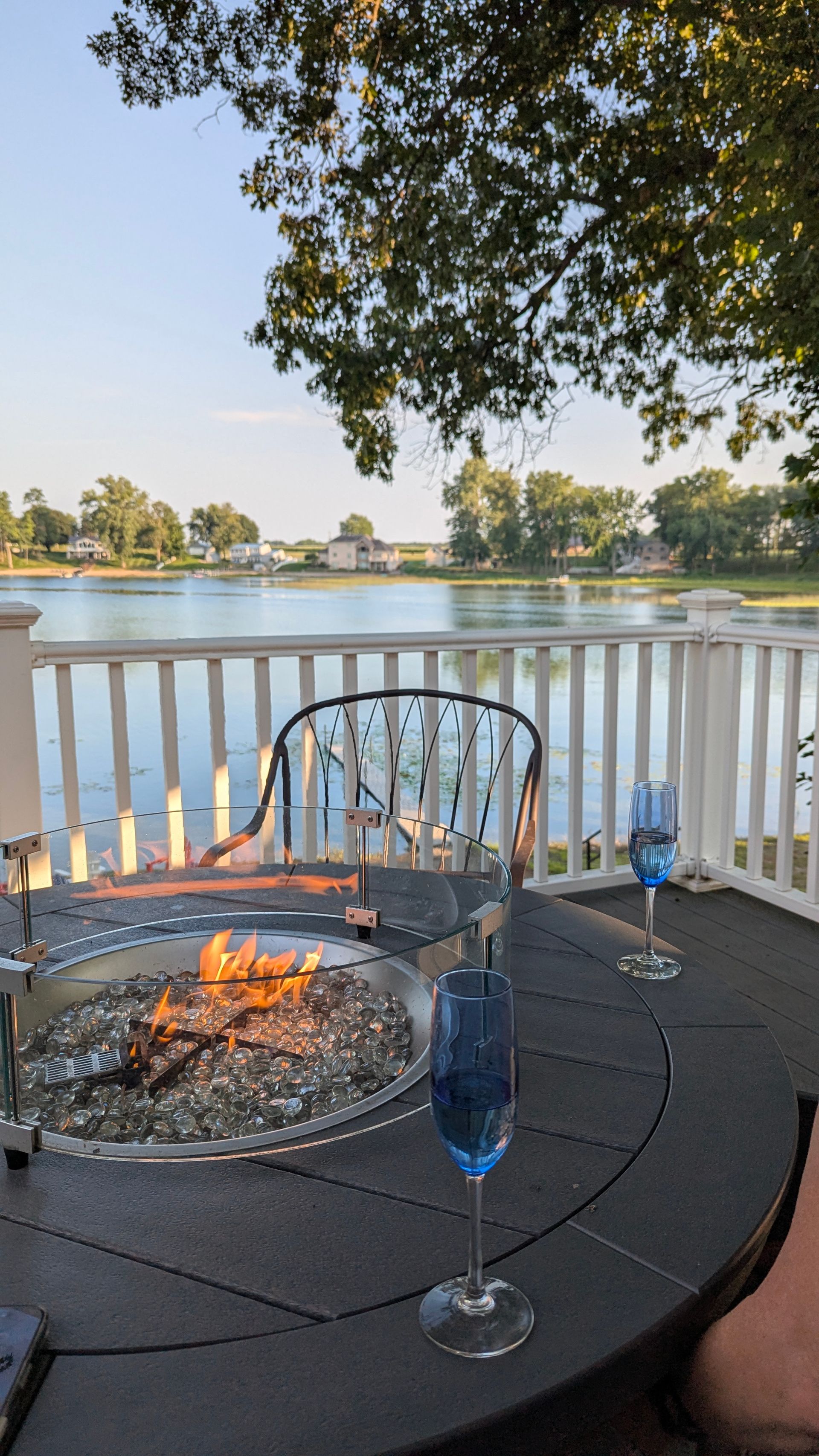 Patio fire pit by a lake at sunset, with a chair, wine glass, and white railing under trees