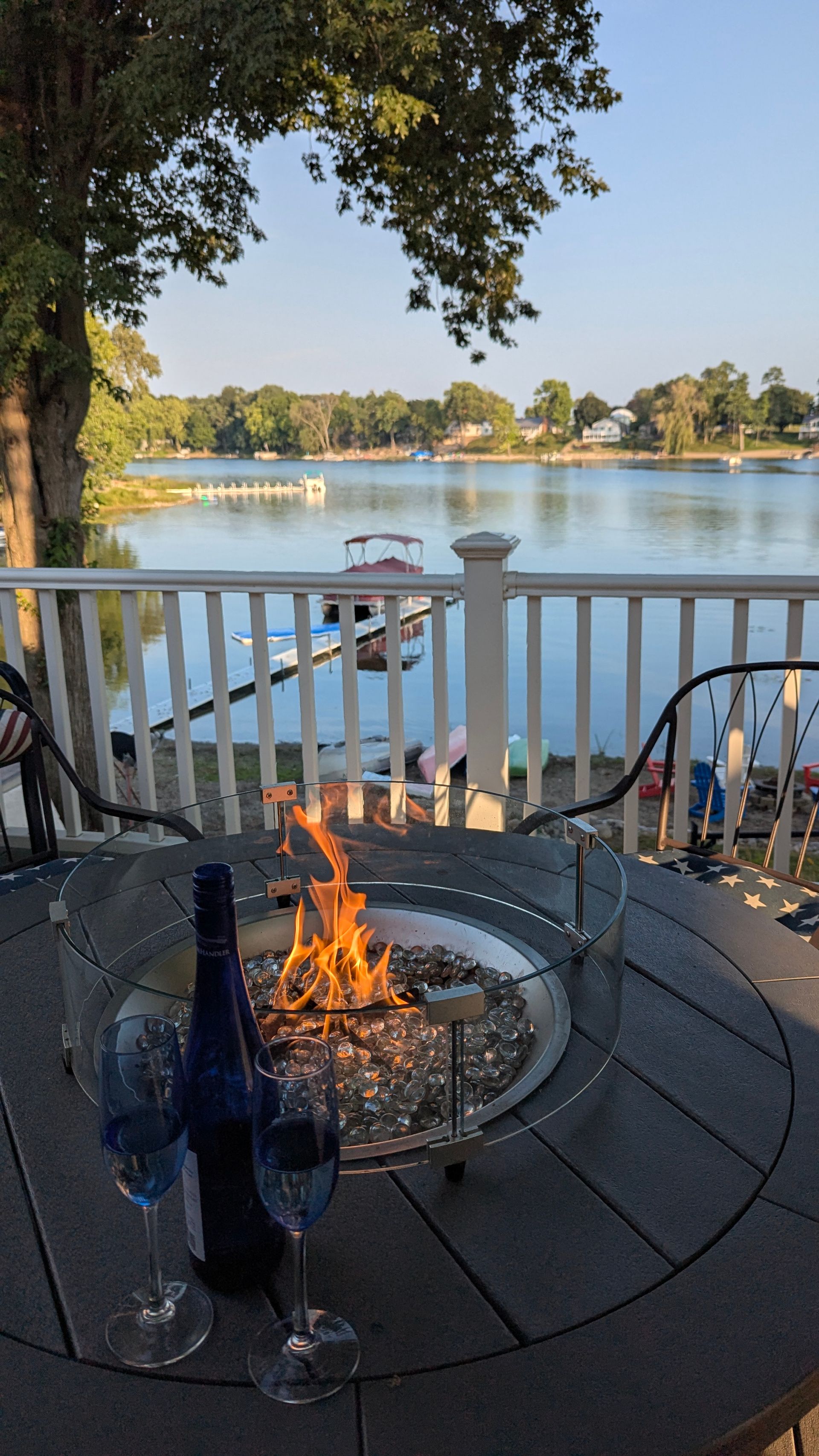 Patio fire pit overlooking a calm lake at sunset, with chairs and a railing in the foreground
