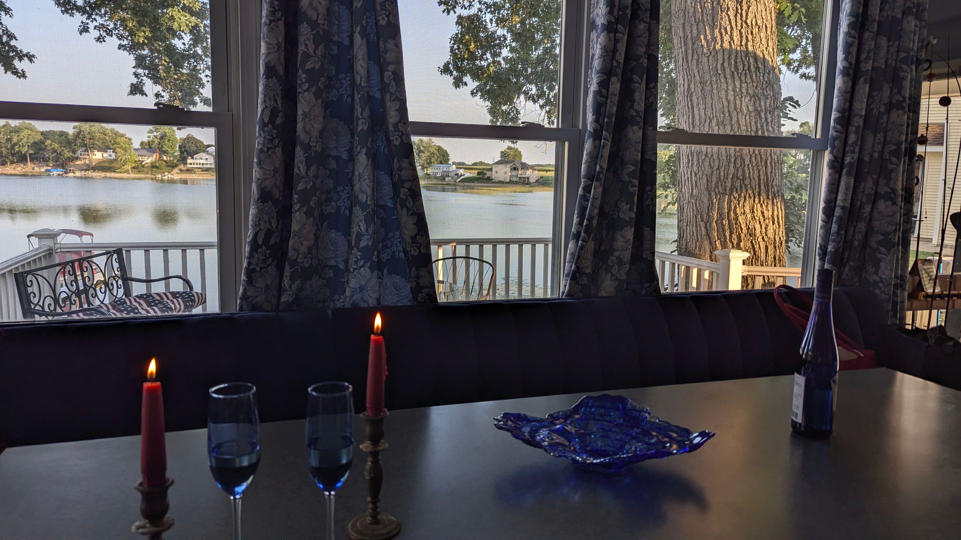 Candlelit table by a lake, with wine glasses and a blue dish in front of large windows.