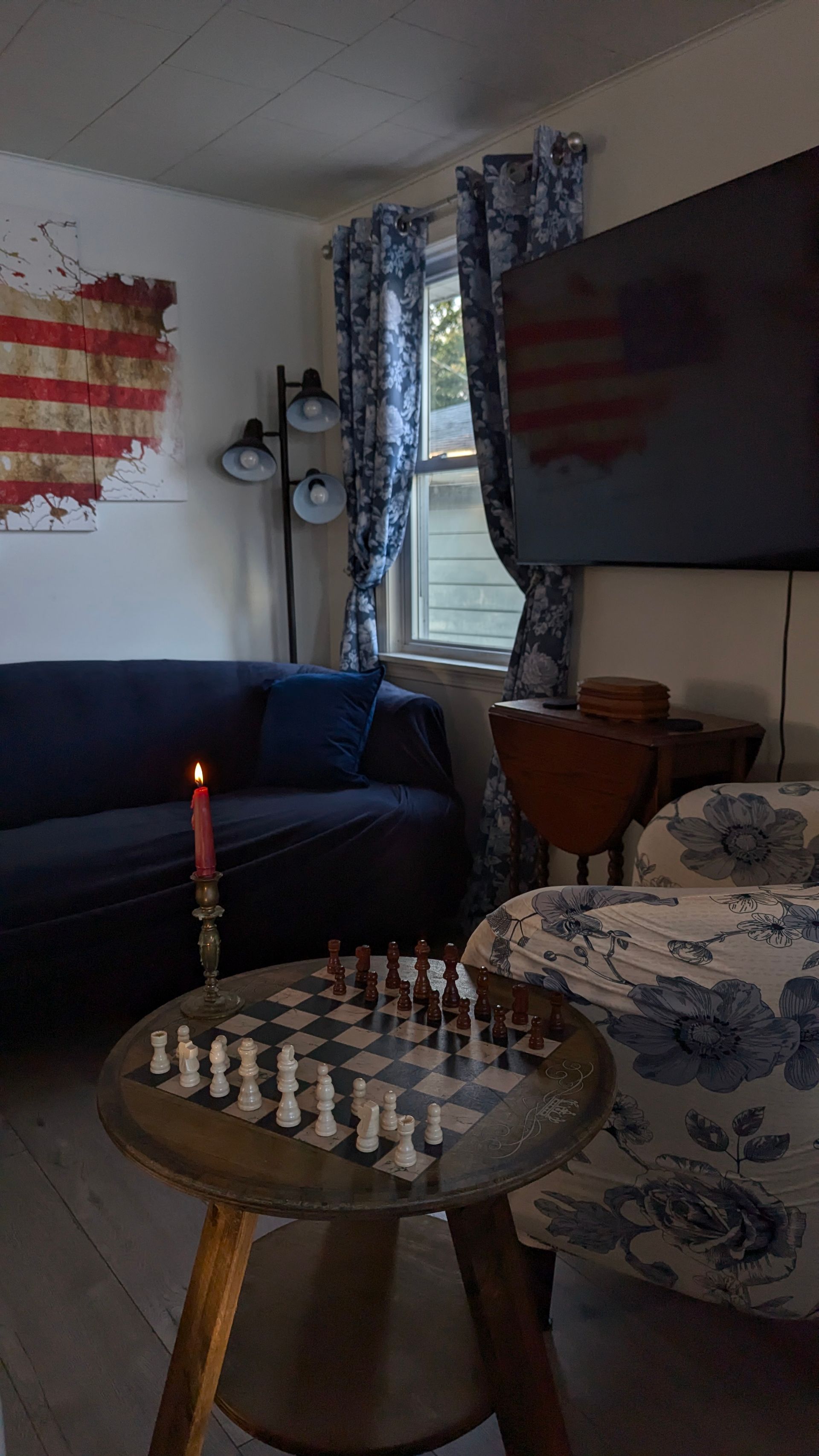 Dim living room with a coffee table full of chess pieces, a lit candle, couch, TV, and window with curtains.