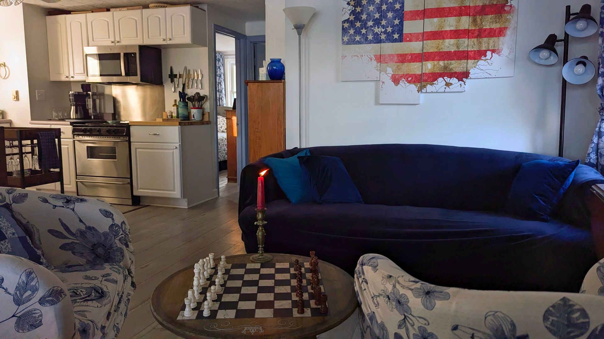 Cozy living room with blue couch, patterned chairs, chessboard table, and kitchen in the background
