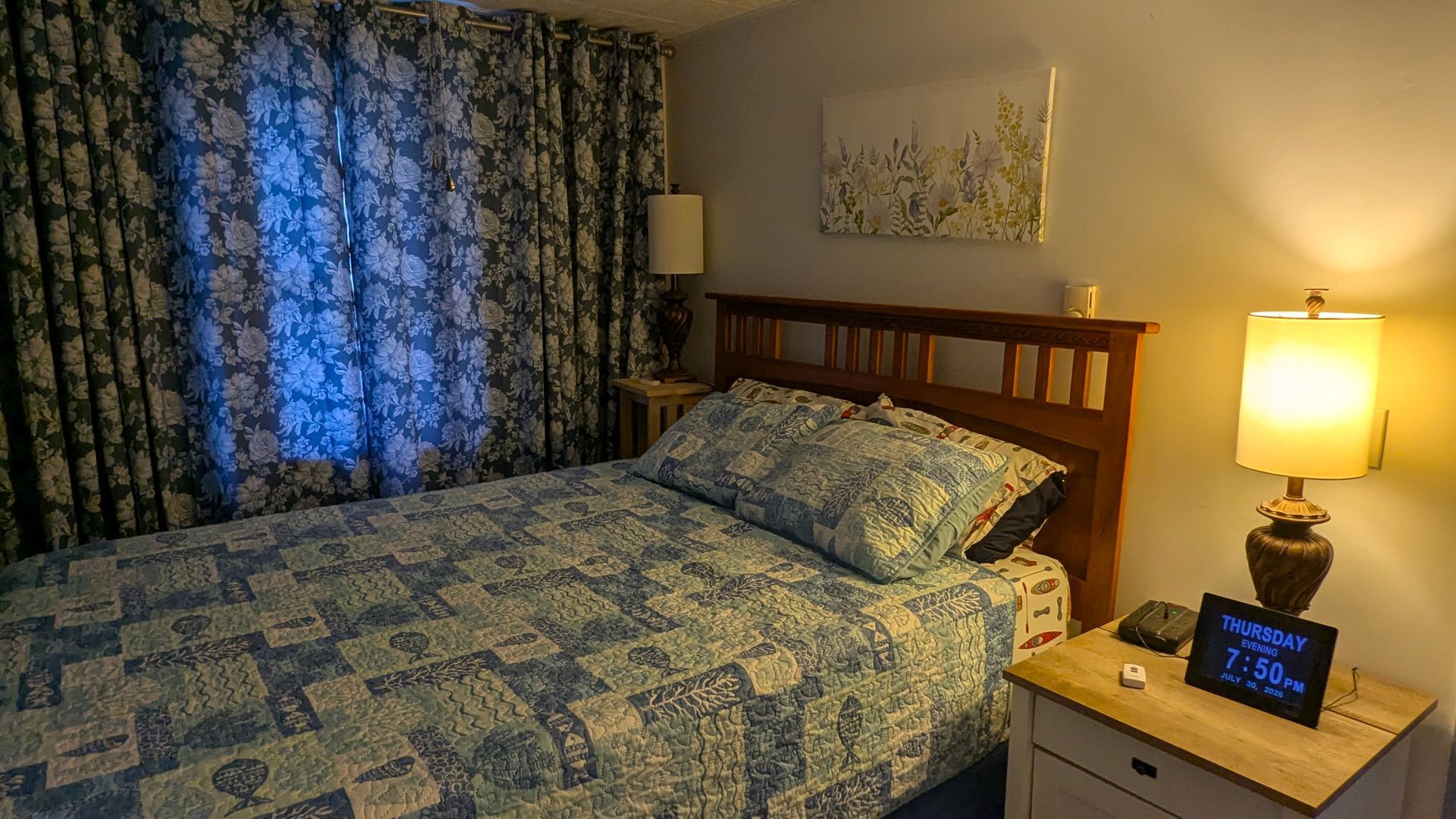 Bedroom with blue patterned bedspread, wooden headboard, lit lamp, and window with dark curtains