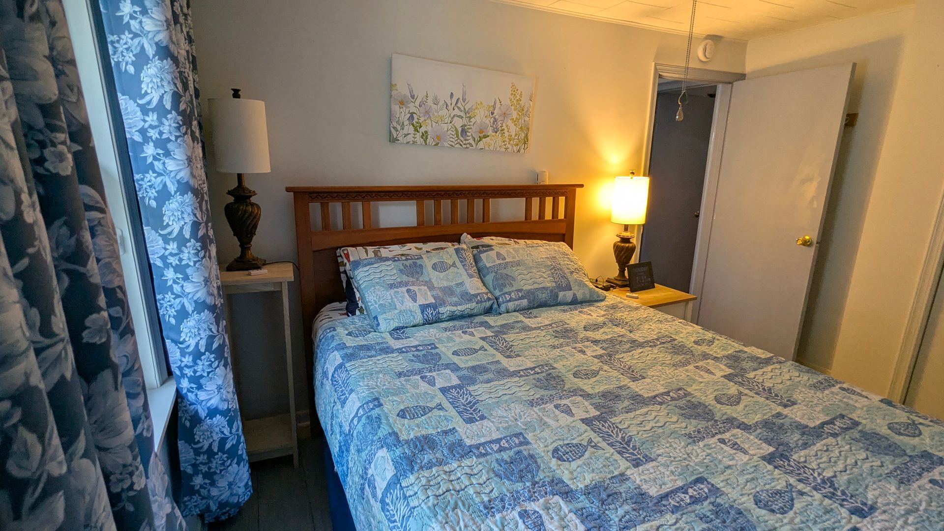 Cozy bedroom with blue patterned bedspread, wooden headboard, bedside lamp, and floral curtains