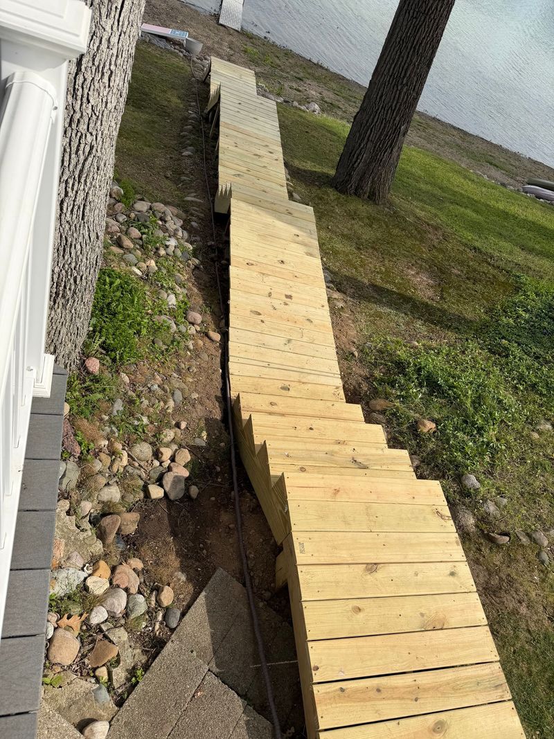 A long wooden walkway beside a house, crossing a muddy trench over grass and rocks.