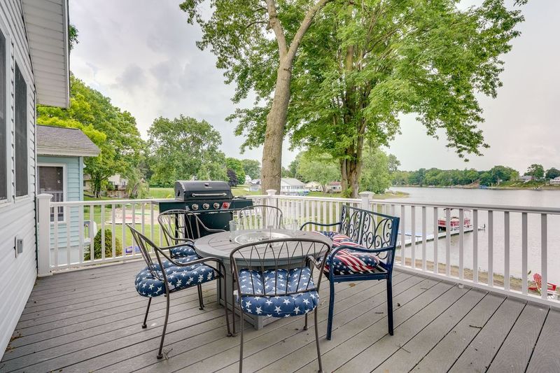 Deck overlooking a lake, with table, chairs, grill, and trees. Overcast sky, white railing.
