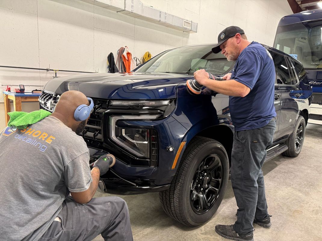 Two people detailing a dark blue SUV, one on the front, one on the hood, in a well-lit shop.