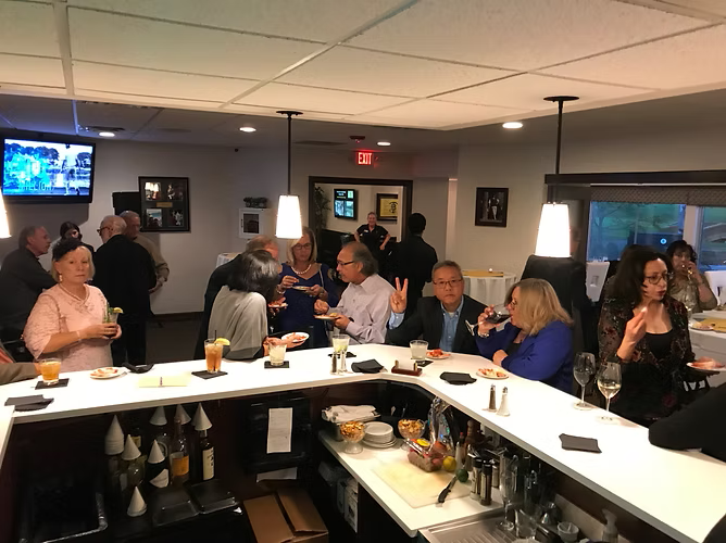 People at a bar, some seated, drinking and talking. Dim lighting, TV on wall, overhead lights.