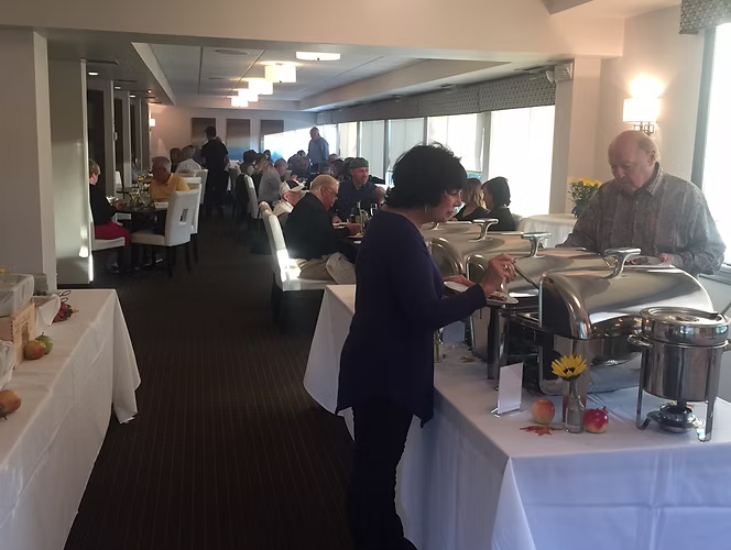 People at a buffet in a brightly lit room. A person serves food. Tables are set for dining.