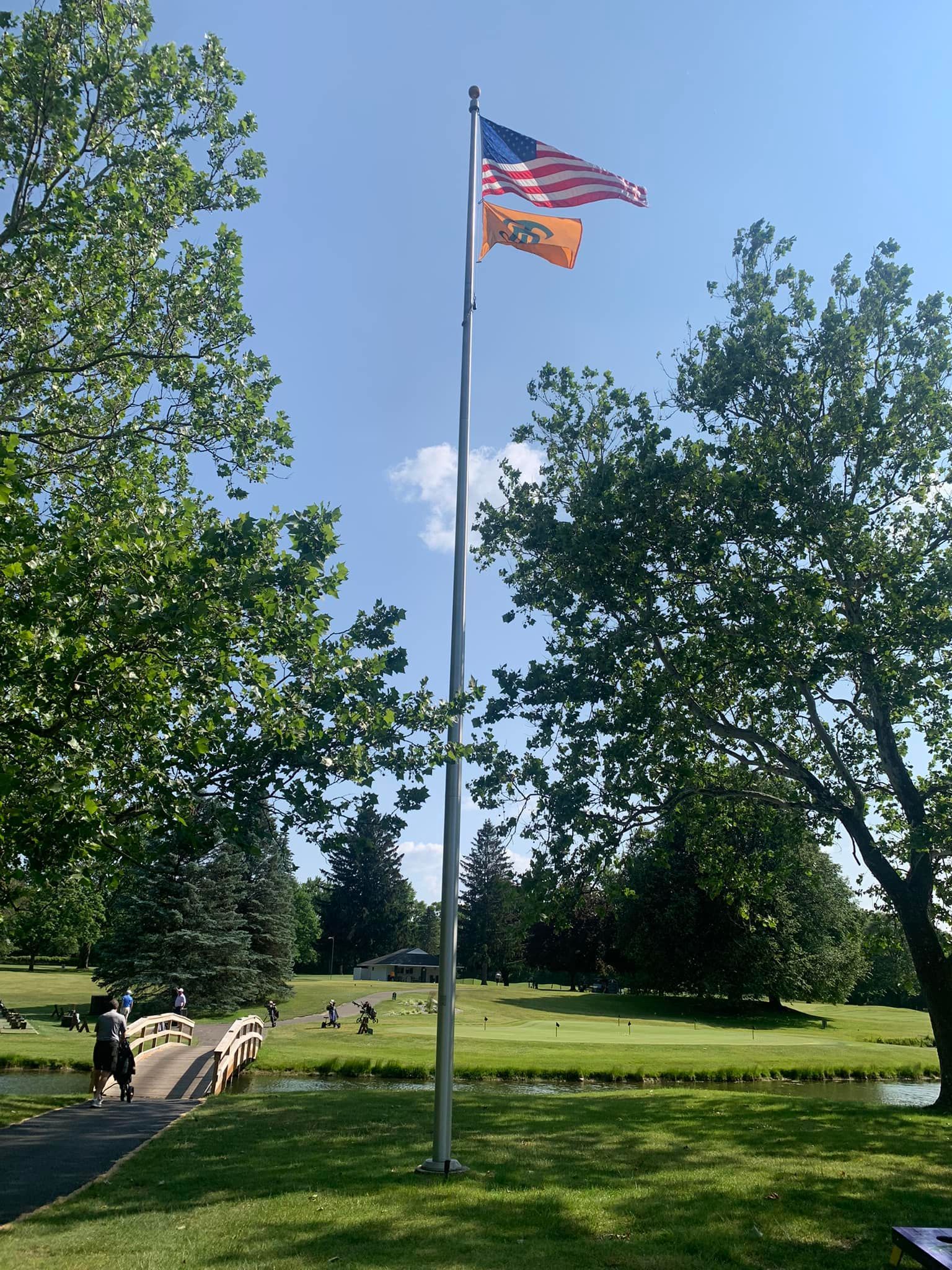 American and orange flags fly on a pole over a golf course with trees and a bridge.