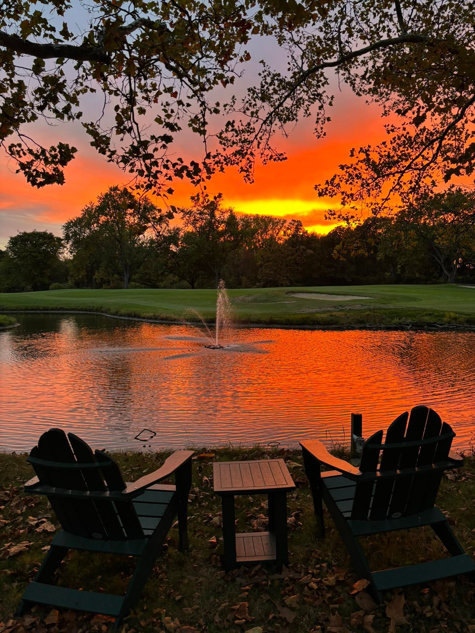 Two Adirondack chairs face a pond reflecting an orange sunset. A small table sits between them.