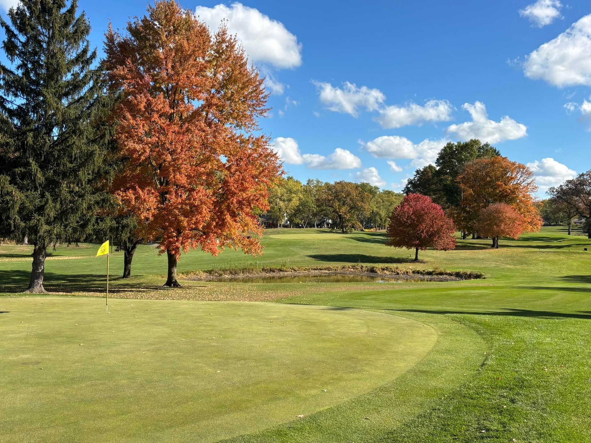 Golf green with fall trees, yellow flag, pond, and blue sky.
