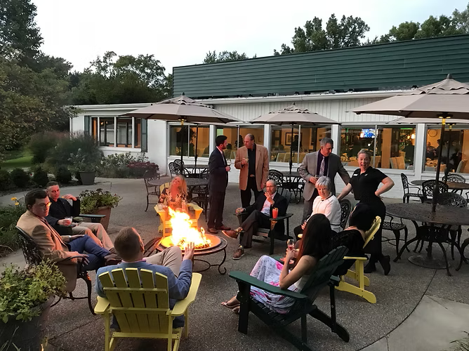 People gathered around a fire pit on a patio outside a building with umbrellas and tables.