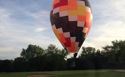 Hot air balloon with orange, yellow, and black checkered pattern floats over a golf course.