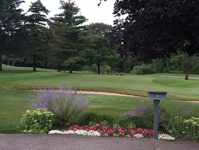 Lush green golf course with bunkers and trees; foreground has colorful flowers, a mailbox, and a paved area.