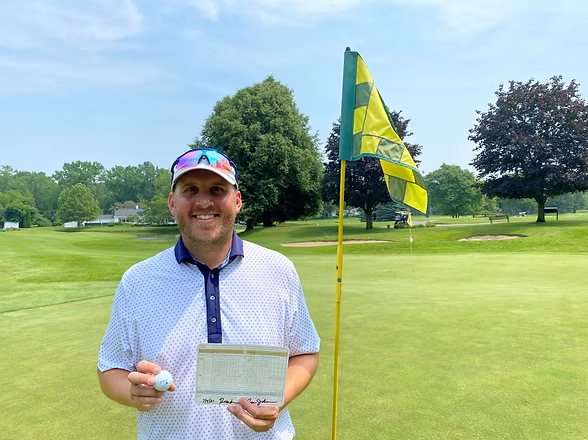 Golfer smiling on a green, holding a scorecard and ball beside a yellow flag on a sunny course