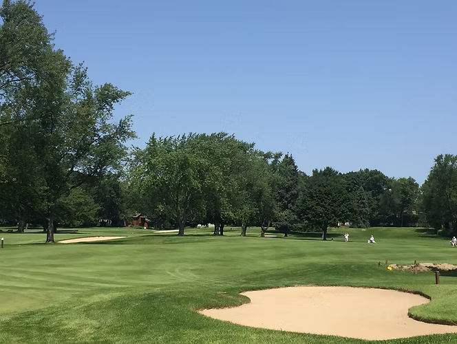 Green golf course with sand traps, trees, and blue sky.