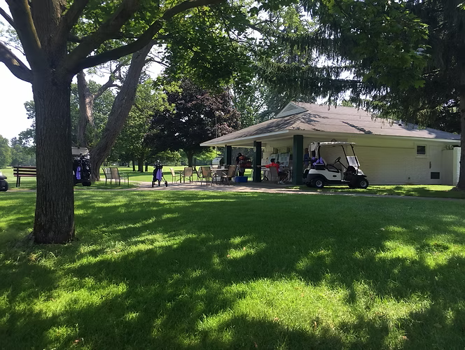 Golf course building with outdoor seating, golf cart, and people on a sunny day.