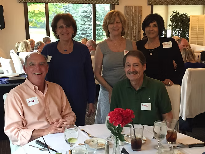 Group of people smiling at a table, some standing behind seated men. Restaurant setting with a window.