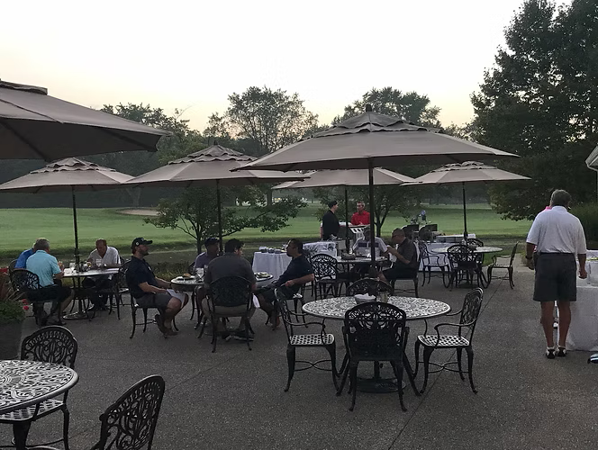 Outdoor patio with tables and umbrellas, people seated, overlooking a golf course.
