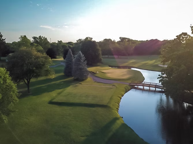 Golf course with bridge over water, leading to a green with trees and sunlight.