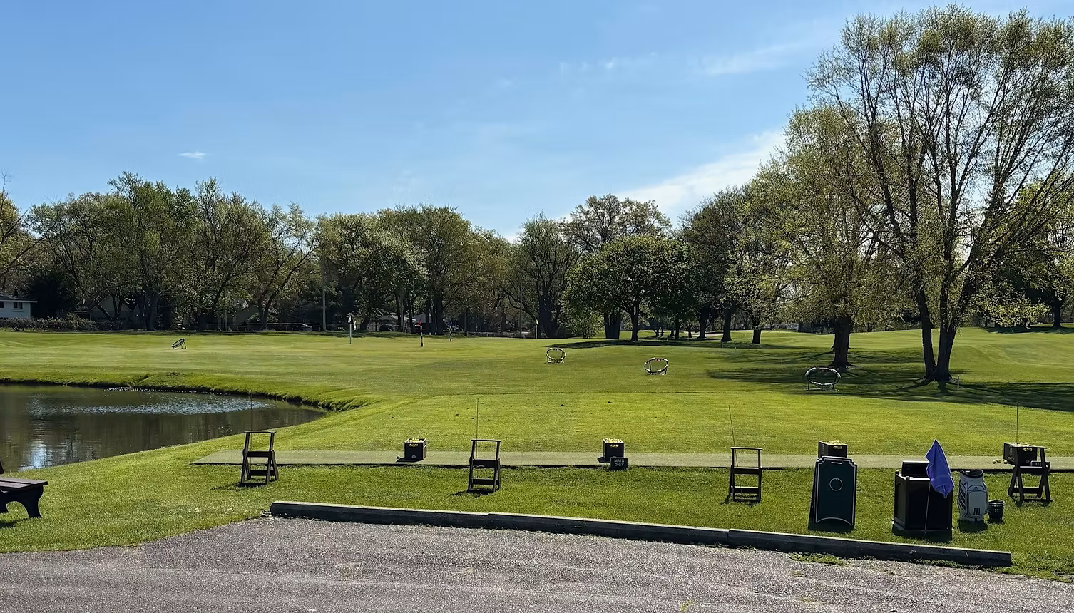 Lush green golf course with trees under a blue sky. A pond is in the foreground.