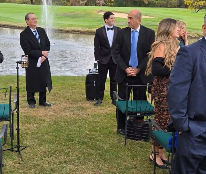 People at an outdoor event with a pond and fountain. Some are standing, others seated in chairs.